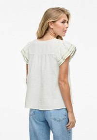 Blusa de algodón gris claro con mangas acampanadas y ribete verde. Presenta un escote en forma de lágrima en la parte posterior y un corte relajado para mayor comodidad. Llevada con jeans azules.