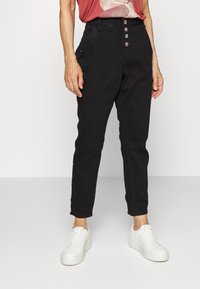 Calças pretas de cintura alta com um corte afunilado, apresentando uma frente com fecho de botão e bolsos laterais, combinadas com ténis brancos.