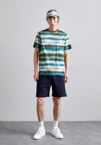 T-shirt à manches courtes avec un motif zigzag multicolore en vert, bleu et blanc, associé à un short marine et des baskets blanches.