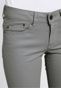 Šedé úzké džíny vyrobené z hladké, elastické tkaniny. Obsahují pět kapes, černé kovové doplňky a viditelné švy.