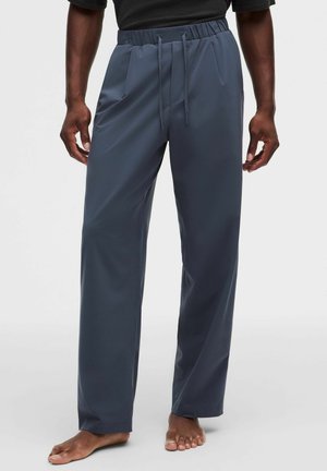 Mann trägt lockere, dunkelgraue Hose mit elastischem Bund und Kordeln, steht barfuß vor einem schlichten hellen Hintergrund.