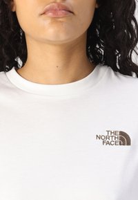 Bijela pamučna majica s okruglim izrezom, koja sadrži smeđi izvezeni logo "The North Face" na lijevoj strani prsa.