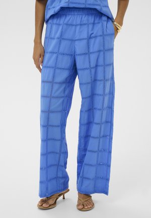 Blaue Hosen mit weitem Bein und quadratischem Lochstickmuster, elastischem Taillenbund, getragen mit beigefarbenen Sandalen mit Absatz und goldenen Armreifen.