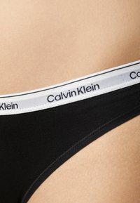 Svart bomulls-thong med en hvit elastisk linning med "Calvin Klein" merkevare og en subtil elastisk tekstur. Nærbilde viser sømdetaljer.