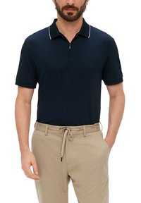 Granatowa koszulka polo z suwakiem, z białym wykończeniem kołnierzyka, wykonana z gładkiego materiału, w połączeniu z beżowymi spodniami ze sznurkiem.