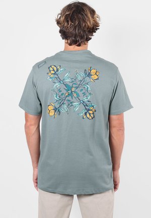 Homme portant un t-shirt vert-gris avec un grand motif floral symétrique jaune et bleu dans le dos, debout sur un fond blanc.