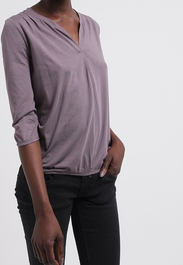Top lilla a scollo a V realizzato in tessuto morbido, con maniche a tre quarti e dettagli di arricciatura sullo scollo, abbinato a jeans neri.
