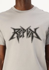 Grå bomulds t-shirt med et sort grafisk logo med skarpe, vinklede bogstaver over brystet. Kortærmet og med en rund halsudskæring.