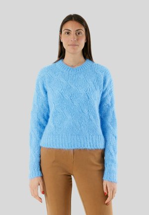 Donna con lunghi capelli castani che indossa un maglione a maglia strutturata azzurro chiaro e pantaloni marroni, in piedi davanti a uno sfondo semplice grigio chiaro.