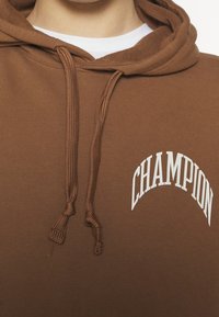 Sudadera con capucha marrón con cordón, con un logo blanco "CHAMPION" en diseño arqueado. Textura suave y tejido acanalado.