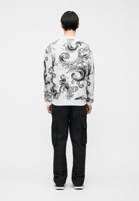 Άτομο που στέκεται με την πλάτη προς τα έξω φορώντας ένα λευκό φούτερ με μαύρα σπειροειδή μοτίβα και μαύρο παντελόνι cargo με πλαϊνές τσέπες.