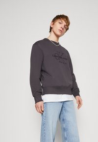 Donkergrijze katoenen sweatshirt met ribgebreide manchetten en zoom, met een zwart Calvin Klein Jeans-logo op de voorkant, gedragen over een wit shirt, gecombineerd met lichtblauwe jeans.