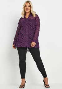 Purple lace tunic with a collar, long sleeves, and an open, airy design. Worn over black skinny jeans and heeled sandals.