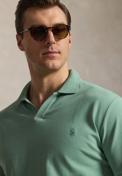 Άνδρας που φοράει γυαλιά ηλίου με φαρδιά καφέ φρύδια και ένα ανοιχτοπράσινο πόλο με ένα μικρό κεντητό λογότυπο, κοιτάζοντας ελαφρώς προς τα πλάγια.