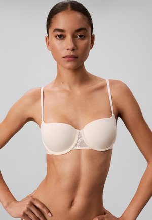 Woman wearing a beige strapless bra with thin adjustable straps and lace detail at the center against a gray background.