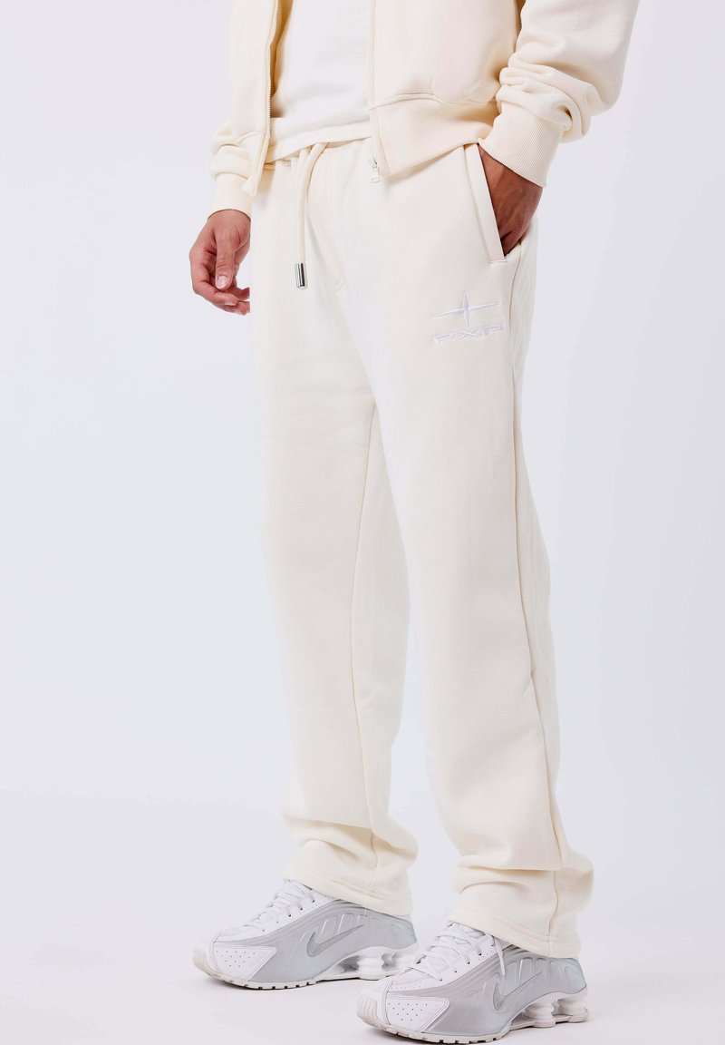 Persona che indossa pantaloni felpati color crema con logo PXP, giacca crema e sneakers Nike bianche e grigie, in piedi con una mano in tasca.