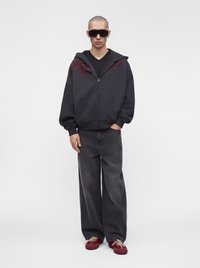 Young man wearing oversized black zip-up hoodie, wide-legged dark jeans, red sneakers, and large black sunglasses standing against white background.