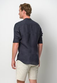 Camicia di lino blu navy con maniche arrotolate, taglio dritto e dettaglio con yoke sul retro, abbinata a pantaloni corti color khaki chiaro.