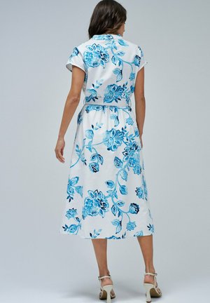 Vestido midi floral con patrones azules sobre tela blanca. Mangas cortas, cintura ajustada y falda fluida. Sandalias planas de tiras beige.
