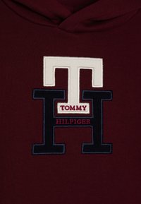 Bordeaux hoodie met een groot geborduurd logo in crème en marineblauw, met de tekst "TOMMY HILFIGER" onder de letter "H."