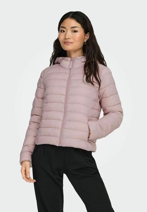 Femme portant une veste matelassée rose clair avec capuche et un pantalon noir, debout avec une main dans la poche devant un fond uni.