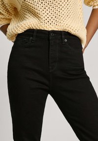 Vaqueros negros de tiro alto confeccionados en denim, con un diseño ajustado, bolsillos frontales y detalles de costura sutiles. Combinados con un suéter de punto.