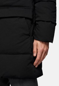 Black puffer coat with textured fabric, featuring a side seam pocket and a fitted cuff. Visible stitching on the surface.