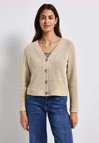 Beige gestrickte Strickjacke mit V-Ausschnitt, langen Ärmeln und vier Knöpfen auf der Vorderseite. Die Textur ist locker mit einem cropped Schnitt. Tragend über einem grauen Oberteil.