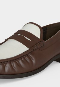 Καφέ και λευκό δερμάτινο μοκασίνι με ραφές μοκασίνου και λεπτομέρεια με λουράκι για κέρμα, εμφανίζεται κοντινά σε ανοιχτό φόντο.