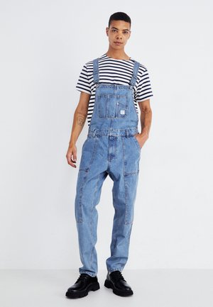 Salopette en denim bleu clair avec bretelles et une poche avant, associée à un t-shirt rayé noir et blanc et des chaussures à lacets noires.