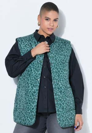 Osoba o krótkich włosach, nosząca złote kolczyki w kształcie koła, turkusową pikowaną kamizelkę w panterkę na czarnej koszuli na guziki oraz szare spodnie.