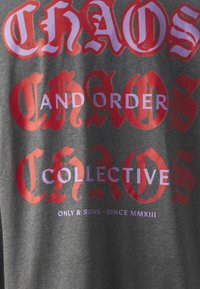 Pilkas marškinėliai su ryškiais raudonais ir violetiniais užrašais: "CHAOS AND ORDER COLLECTIVE", su stilingu šriftu ir matomu audinio tekstūra.