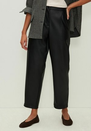 Frau trägt hochtaillierte schwarze, weite Lederhosen, braune Wildleder-Schuhe, ein weißes Oberteil und eine Jacke mit Hahnentrittmuster.