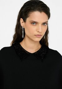 Black sweater with a textured, shiny black collar. The model also wears long silver chain earrings. Background is plain white.