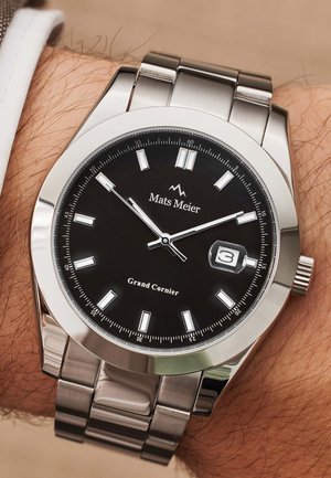 Montre-bracelet en acier inoxydable avec un cadran noir, index des heures en argent, fenêtrage de date et bracelet poli. Présente un branding distinctif et une couronne texturée.