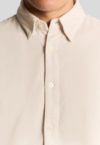 Lyse beige knap-up skjorte med ribbet tekstur og en foldet krave. Har fire forreste knapper og en ren finish langs knappenummeret.