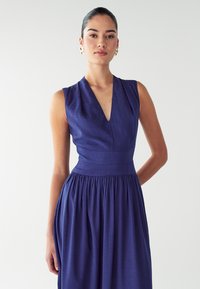 Mornarici plava haljina bez rukava s V-izrezom, krojena u struku, s nabranoj suknjom. Izrađena od strukturirane tkanine, s minimalističkim dizajnom.
