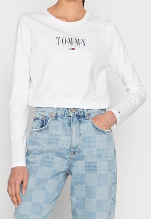 Vrouw draagt een wit Tommy Jeans shirt met lange mouwen en logo op de borst, met haar hand in de zak van lichtblauwe geruite hoge taille jeans.