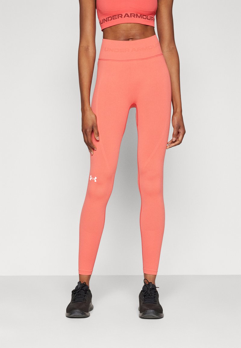 Nő korall színű, Under Armour magas derékú leggingsben és hozzáillő sportmelltartóban, fekete sportcipővel, egyszínű háttér előtt állva.