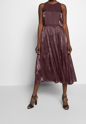 Femme portant une robe midi sans manches, brillante, couleur prune, avec une jupe plissée et des sandales noires à talons hauts, debout sur un fond blanc.
