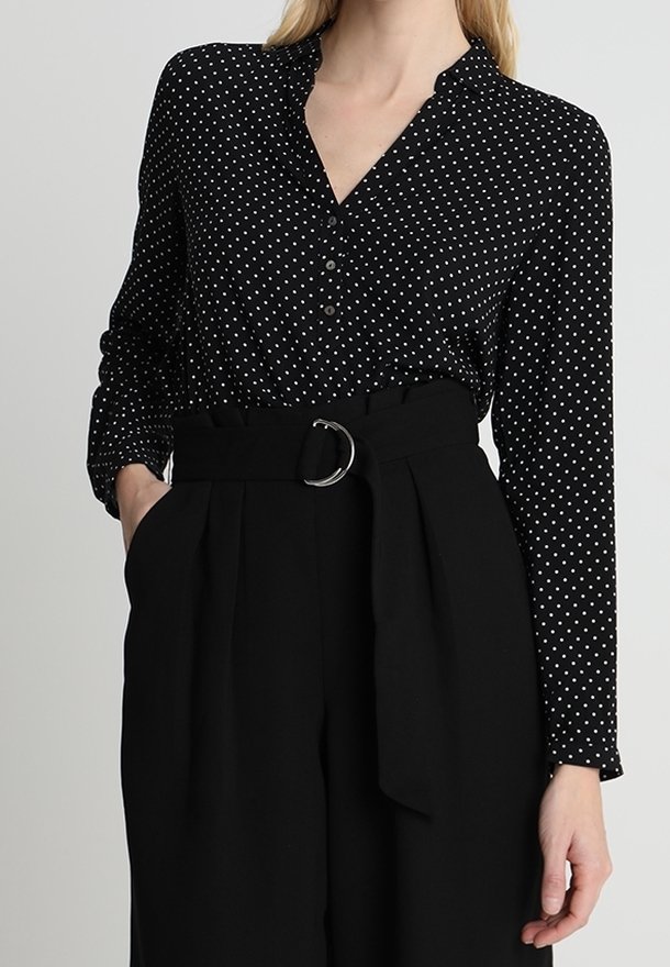 Blouse noire à pois avec patte de boutonnage et col, associée à un pantalon noir taille haute, doté d'une ceinture avec une boucle circulaire.