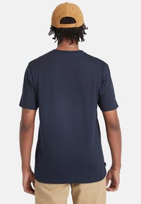Námorně modré tričko s krátkými rukávy a kulatým výstřihem. Měkká bavlněná tkanina, běžný střih. Nosené s hnědou kordovou čepicí, zobrazeno z pohledu zezadu.