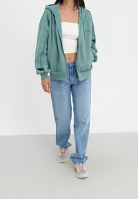 Giovane donna che indossa un'ampia felpa teal con zip, una canotta bianca, jeans larghi di un azzurro chiaro e scarpe sportive beige, in piedi contro uno sfondo bianco.