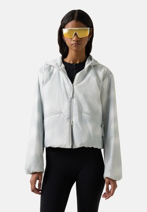 Kvinde iført en lysegrå hættetrøje med lynlås, sorte leggings og store gule, futuristiske solbriller, stående mod en ensfarvet baggrund.