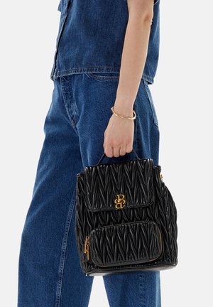 Mano sosteniendo un bolso de cuero negro acolchado con logotipo dorado y bolsillo con cremallera, vistiendo jeans de mezclilla azul y top sin mangas con pulsera dorada.