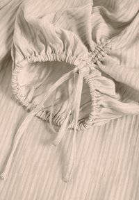 Beige Stoff mit gerafften elastischen Bündchen, die mit dünnen Stoffkordeln gebunden sind, zeigt detaillierte Nähte und eine weiche Textur.