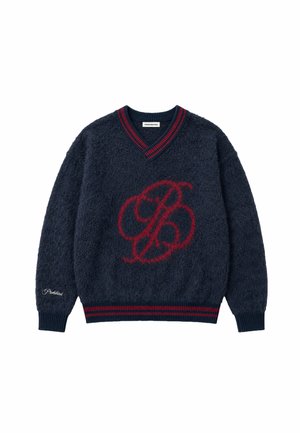 Homme portant un pull bleu marine avec un motif rouge, un bonnet rouge tricoté, tenant un manteau à carreaux marron sur son épaule devant un fond clair uni.