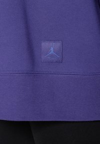 La tela púrpura presenta un acabado texturizado, con un parche rectangular del logo azul de Jordan en la parte inferior, acentuado por material negro en la parte inferior.