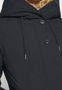 Mustat ulkoilutakit, joissa on korkea kaulus, kaksi nappia, kiristysnauhoilla varustettu huppu ja sileä kangas. Minimalistinen muotoilu, jossa on toiminnallisia yksityiskohtia.