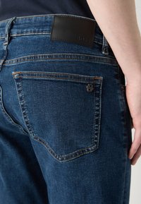 Jeans din denim de culoare albastru închis, cu o textură netedă, având un patch din piele neagră cu logo pe banda din spate și un mic accent metalic aproape de buzunar.